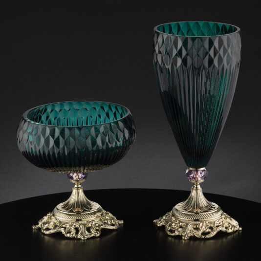 A pair of elegant diamond-faceted green glass pieces with intricate patterns. The shorter, bowl-shaped vessel is on the left and the taller, vase-shaped piece is on the right. Both items have ornate gold bases with detailed design work, set against a dark background.