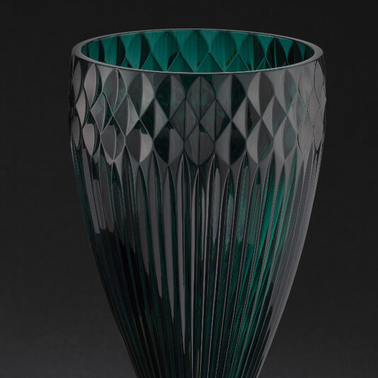 Diamond Faceted Green Glass Vase And Bowl A close-up of the Diamond Faceted Green Glass Vase And Bowl against a black background. The vase showcases an intricately patterned upper portion with diamond shapes and vertical grooves on the lower half, capturing light to create contrasting highlights and shadows.