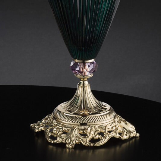Diamond Faceted Green Glass Vase And Bowl A close-up of an ornate, gold-colored lamp base with intricate floral detailing. The lamp base supports a dark green, vertically ridged glass shade reminiscent of the Diamond Faceted Green Glass Vase And Bowl collection. There is a purple, faceted gemstone accent between the shade and the base against a black background.