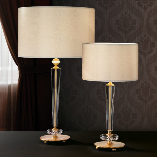 Two Elegant Glass Table Lamps with beige fabric shades and crystal bases stand on a black surface in front of dark, patterned wallpaper. The taller lamp is positioned on the left, while the shorter one sits on the right. A window with brown curtains is visible in the background.