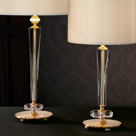 Elegant Glass Table Lamp Two Elegant Glass Table Lamps, featuring tall transparent bodies and cylindrical shades, sit on a dark surface. The lamps boast gold-colored bases with intricate detailing, beautifully illuminated against a dark, patterned background.