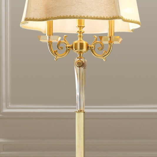 Luxury Classical Floor Lamp The Luxury Classical Floor Lamp features a gold and transparent glass base adorned with intricate floral detailing, topped with a beige fabric lampshade that includes a decorative border. The lamp stands against a beige wall accented by white wainscoting.