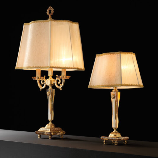 Two elegant High End Amber Glass Table Lamps with beige, pleated lampshades set against a dark background. The left lamp, a premium model with a taller, ornate base and candelabra-style arms, contrasts sharply with the right's shorter and simpler base. Both models boast gold accents and intricate designs that add an air of sophistication.