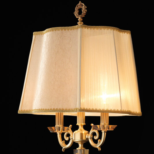 High End Amber Glass Table Lamp The High End Amber Glass Table Lamp is a vintage-style brass lamp with a rectangular beige fabric lampshade. It boasts elaborate detailing on its base, a decorative finial atop the shade, and an amber glass accent that casts warm, soft light against a dark background.