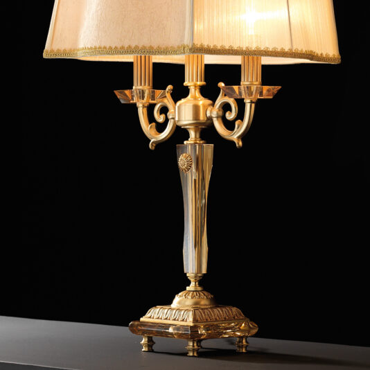 High End Amber Glass Table Lamp The High End Amber Glass Table Lamp is an elegant piece with ornate detailing. It features a beige fabric lampshade and a transparent, tapered amber glass central column. Multiple decorative arms hold lightbulbs, creating a warm, inviting glow. The intricately designed base adds to its high-end appeal.
