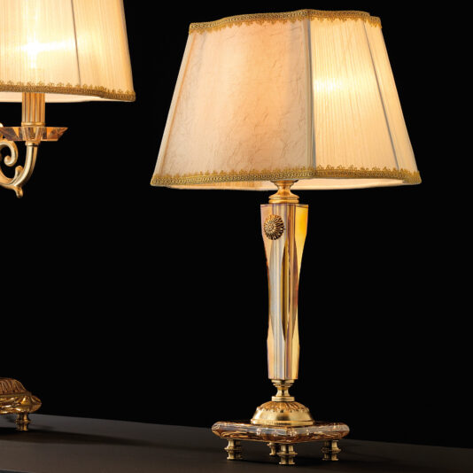 High End Amber Glass Table Lamp A **High End Amber Glass Table Lamp** featuring a textured square cream lampshade, ornate detailing, and a sturdy base is turned on, casting warm light in its surroundings against a dark background. On the left, another similar lamp is partially visible.