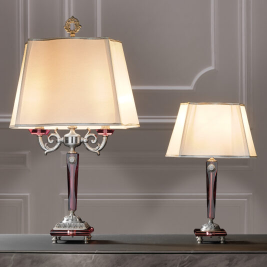 A photo depicts two exquisite High End Antique Rose Glass Table Lamps with beige shades resting on a marble surface. The larger, luxurious lamp showcases an antique rose glass base adorned with red and silver accents and intricate detailing. The smaller lamp presents a more straightforward design while maintaining the same elegant color scheme. Both lamps are set against a refined grey wall, enhancing their sophisticated appeal.
