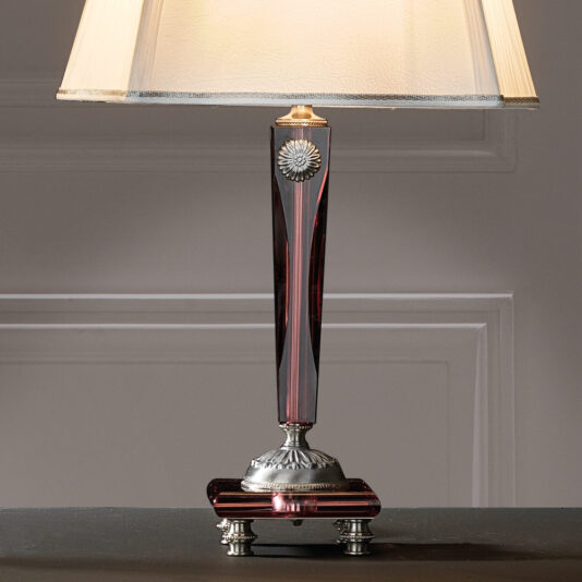 High End Antique Rose Glass Table Lamp The High End Antique Rose Glass Table Lamp features a rectangular, dark red transparent base adorned with ornate silver accents. It stands on a small platform supported by four feet. The classic beige lampshade slightly flares upwards, all set against a simple, white paneled wall.