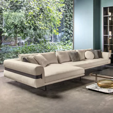 High End Contemporary Modular Corner Sofa A modern living room featuring the High End Contemporary Modular Corner Sofa in beige adorned with multiple cushions. The space is illuminated by floor-to-ceiling windows that reveal a view of lush greenery outside. In the center, there's a coffee table with a marble top, and against a teal wall in the background stands a bookshelf.
