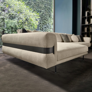 High End Contemporary Modular Corner Sofa A **High End Contemporary Modular Corner Sofa** in beige with a black accent strap along the back is set on a dark carpeted floor in front of a large window that offers a lush green garden view. The room features dark walls and shelves, and various cushions are arranged on the sofa.
