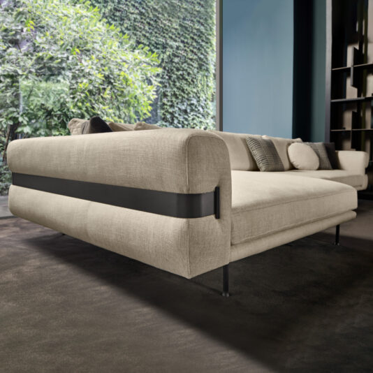 A **High End Contemporary Modular Corner Sofa** in beige with a black accent strap along the back is set on a dark carpeted floor in front of a large window that offers a lush green garden view. The room features dark walls and shelves, and various cushions are arranged on the sofa.