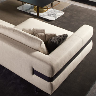High End Contemporary Modular Corner Sofa A modern living room features a High End Contemporary Modular Corner Sofa adorned with several cushions, including a black one. The sofa is positioned on a dark carpet, and in the background, a coffee table with a marble top and a gold bowl can be seen. The overall decor is contemporary.