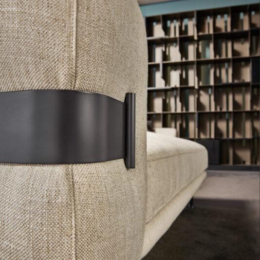 A close-up of the back of a High End Contemporary Modular Corner Sofa, featuring a dark metal bracket. The beige couch is placed in front of a geometric bookshelf adorned with various items, all illuminated by soft, natural light. In the background, there's a blue wall and an area with a dark floor.