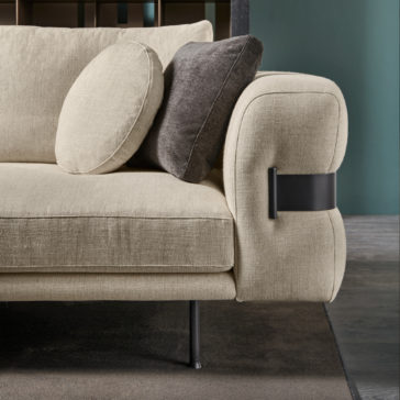 High End Contemporary Modular Corner Sofa Close-up of the High End Contemporary Modular Corner Sofa in beige, featuring rounded armrests and metal accents, adorned with two cushions – one beige and one dark gray. The sofa is positioned on a dark rug against a greenish-blue wall. In the background, there is a wood and metal partition.