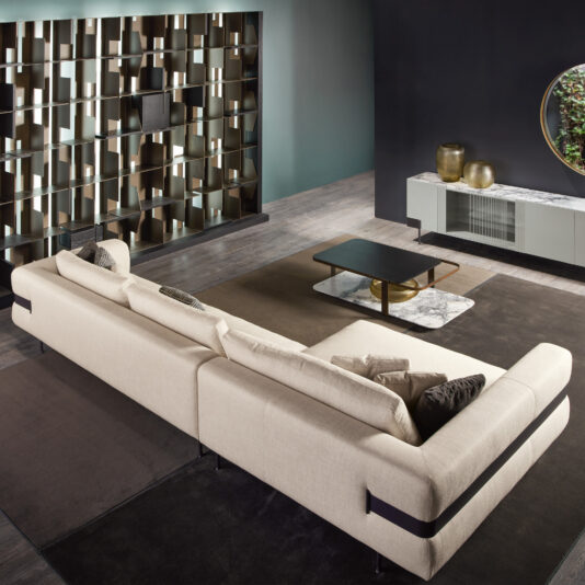 A modern living room featuring a High End Contemporary Modular Corner Sofa, a dark wood coffee table, and a sleek entertainment unit. A decorative shelving unit spans the back wall, with a round mirror hanging above. The space is adorned with minimalist decor in neutral tones.