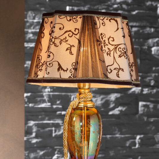 A High End Ornate Amber Glass Table Lamp stands against a textured gray stone wall. The lampshade features a floral pattern with brown and gold tones, complemented by a golden cord wrapped around the lamp's neck.