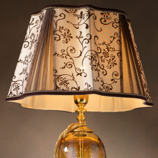 Introducing the High End Ornate Amber Glass Table Lamp, a luxurious piece featuring a golden base and a cream-colored lampshade adorned with intricate black floral designs and dark trim. The lamp's sophisticated base is highlighted by an amber glass accent intertwined with a golden rope, enhancing its elegant aesthetic.