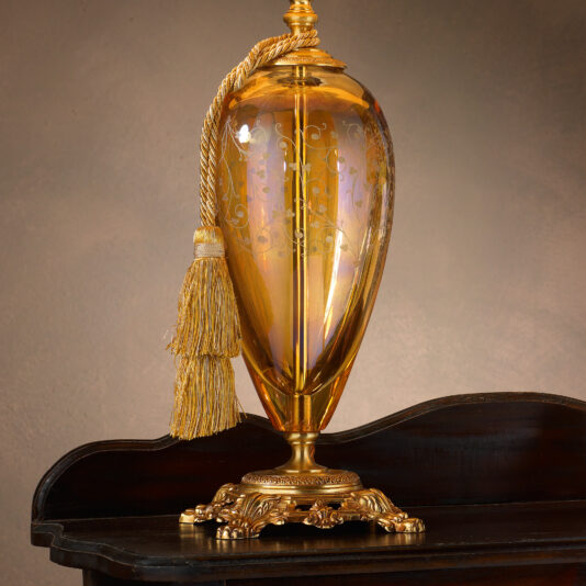 The High End Ornate Amber Glass Table Lamp, with its intricate golden detailing and matching gold tassel, is placed on a dark wooden table. Featuring a decorative base and an ornate design, the lamp creates an elegant and classic aesthetic against a subtle beige background.