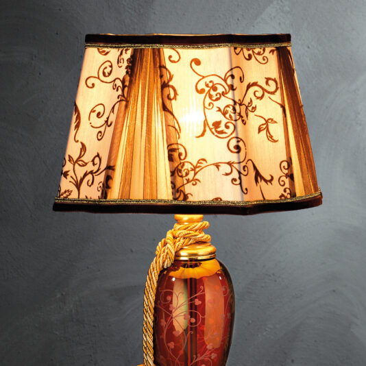 The High End Ornate Ruby Glass Table Lamp showcases a sophisticated ruby glass body paired with a reddish-brown base adorned with an intricate design. Topping the lamp is a decorated beige lampshade that features an elaborate floral pattern, while a gold rope accentuates the upper part of the base, enhancing its decorative charm. The lamp stands against a textured gray wall background, further highlighting its elegant aesthetic.
