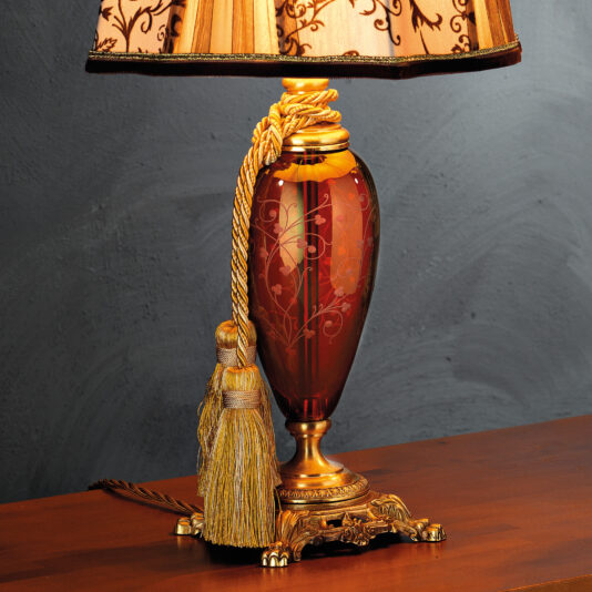 The High End Ornate Ruby Glass Table Lamp is an exquisite decorative piece featuring an intricately engraved ruby glass base with floral designs. It boasts a golden tassel accent and is topped with a beige and gold patterned lampshade. The lamp elegantly rests on a polished wooden surface set against a gray background.