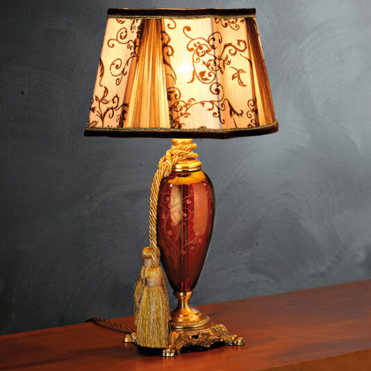 The High End Ornate Ruby Glass Table Lamp features a ruby glass base adorned with intricate patterns and a gold rope accent with a tassel. The beige lampshade, decorated with floral designs and brown trim, sits elegantly on a wooden surface against a grey wall.