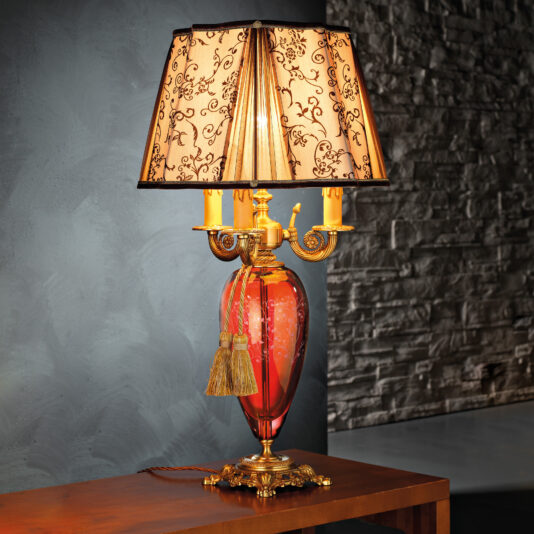 The High End Ornate Ruby Glass Table Lamp sits on a wooden surface, boasting a decorative ruby glass body, golden accents, and two beige tassels. Its ornate base adds to the vintage charm, while the elegantly floral-patterned lampshade casts a warm glow. The background features a textured stone wall.