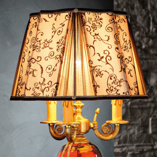 A close-up of the High End Ornate Ruby Glass Table Lamp, featuring an intricate, square beige lampshade adorned with black floral patterns. The lamp boasts a gold-colored, candelabra-style base with elegantly curved arms and candle-like light bulbs. The backdrop showcases a textured gray wall.