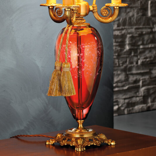 The High End Ornate Ruby Glass Table Lamp features an intricately designed ruby glass vase base adorned with golden floral patterns and tassels. The lamp itself showcases elegant, ornate golden details and rests on a decorative gold base. It is perfectly set against a textured gray wall on a wooden surface.