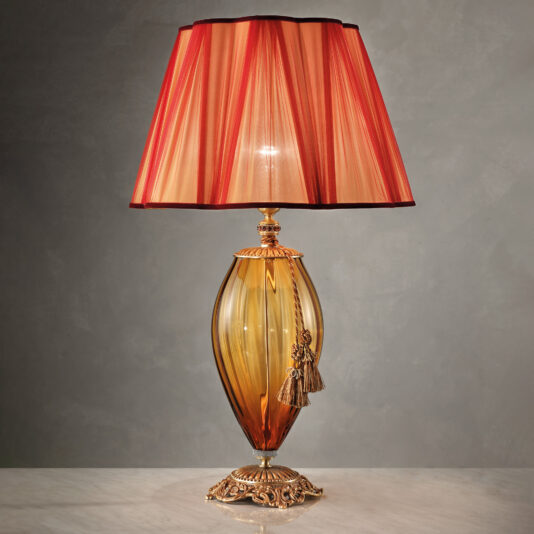 The Large Amber Blown Glass Table Lamp is an elegant piece with a gold-colored amber blown glass base and a red and gold striped fabric lampshade. It boasts intricate gold detailing on the base and is adorned with a decorative gold tassel hanging from the fixture, all set on a marble surface.