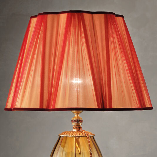 Large Amber Blown Glass Table Lamp A close-up image of the Large Amber Blown Glass Table Lamp, showcasing its ornate gold base with intricate decorative details and a pleated lamp shade. The shade is predominantly red with orange hues, and it emits a warm, ambient glow against the neutral gray wall surface.