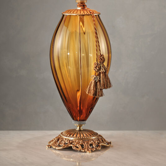 Large Amber Blown Glass Table Lamp The Large Amber Blown Glass Table Lamp, featuring a rounded, elongated shape and resting on a marble surface, showcases a decorative gold metal base with intricate detailing and a tassel attached to the top portion. The background is set against a plain, neutral-colored wall.