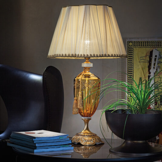 A lit, elegant Large Amber Glass Table Lamp With Swarovski Crystals and a pleated, beige lampshade sits on a glass table next to a stack of books and a potted plant with long, slender green leaves. The background features a dark chair and a framed piece of art.