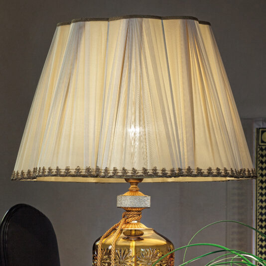 The Large Amber Glass Table Lamp With Swarovski Crystals features a golden base embellished with intricate designs and is topped with a sheer white lampshade accented by delicate trim. The large amber glass incorporates Swarovski crystals, casting a warm glow. In the background, there's a plant along with part of a framed picture on the wall.
