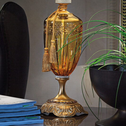 A Large Amber Glass Table Lamp With Swarovski Crystals, featuring an intricate etched pattern and ornate designs, sits on a desk with two tassels hanging from its neck. To the right, there is a black bowl holding a plant with long, slender green leaves. On the left side of the desk, there's a stack of blue folders.