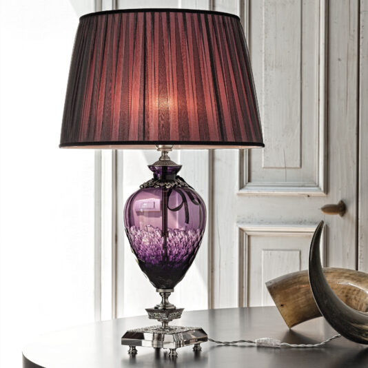 A Large Amethyst Cut Glass Table Lamp with a pleated purple lampshade sits on a dark wooden table. Behind the lamp is a rustic white wooden door, and part of a horn decoration is visible on the table.