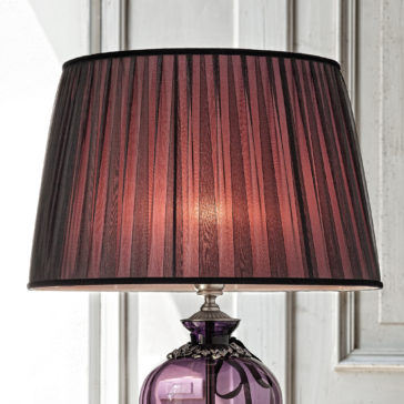 Large Amethyst Cut Glass Table Lamp The Large Amethyst Cut Glass Table Lamp features a pleated, dark purple lampshade and a shiny, purple glass base adorned with a small decorative chain near the top. It is set against a white paneled background.
