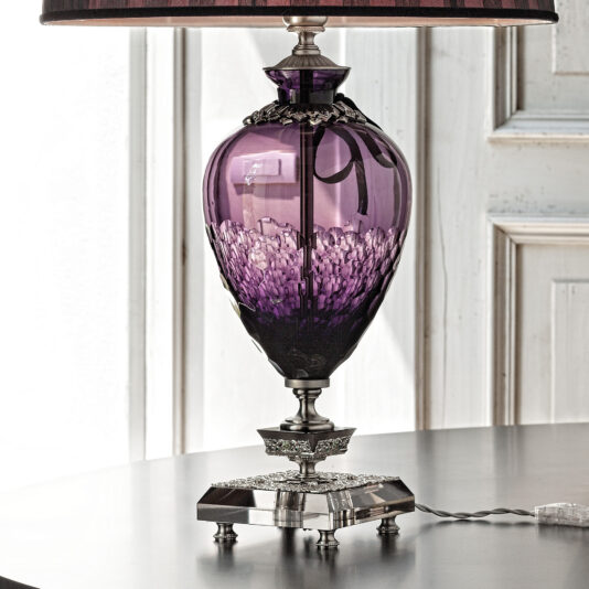 The Large Amethyst Cut Glass Table Lamp boasts a beautiful dark purple base adorned with ornate silver embellishments. It is mounted on a metal pedestal featuring intricate detailing, and the partially visible lampshade at the top enhances its elegance. Positioned on a light-colored table, this lamp adds an elegant touch to any room.