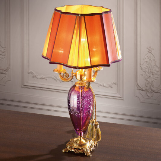 The Large Candelabra Style Glass Table Lamp features a purple glass base etched with intricate white patterns and adorned with gold detailing and elegant hanging tassels. Its warm orange pleated lampshade emits a soft, inviting glow. Perfectly positioned on a wooden surface, this lamp stands out beautifully against an ornate, pale wall.