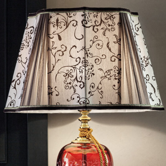 Large Luxury Ornate Ruby Glass Table Lamp The Large Luxury Ornate Ruby Glass Table Lamp boasts a ruby glass base with intricate design details, complemented by a light beige lampshade adorned with delicate dark floral patterns. Its gold-toned fitting and rope detail near the top of the base add to its elegant presence, making it stand out gracefully against any neutral background.