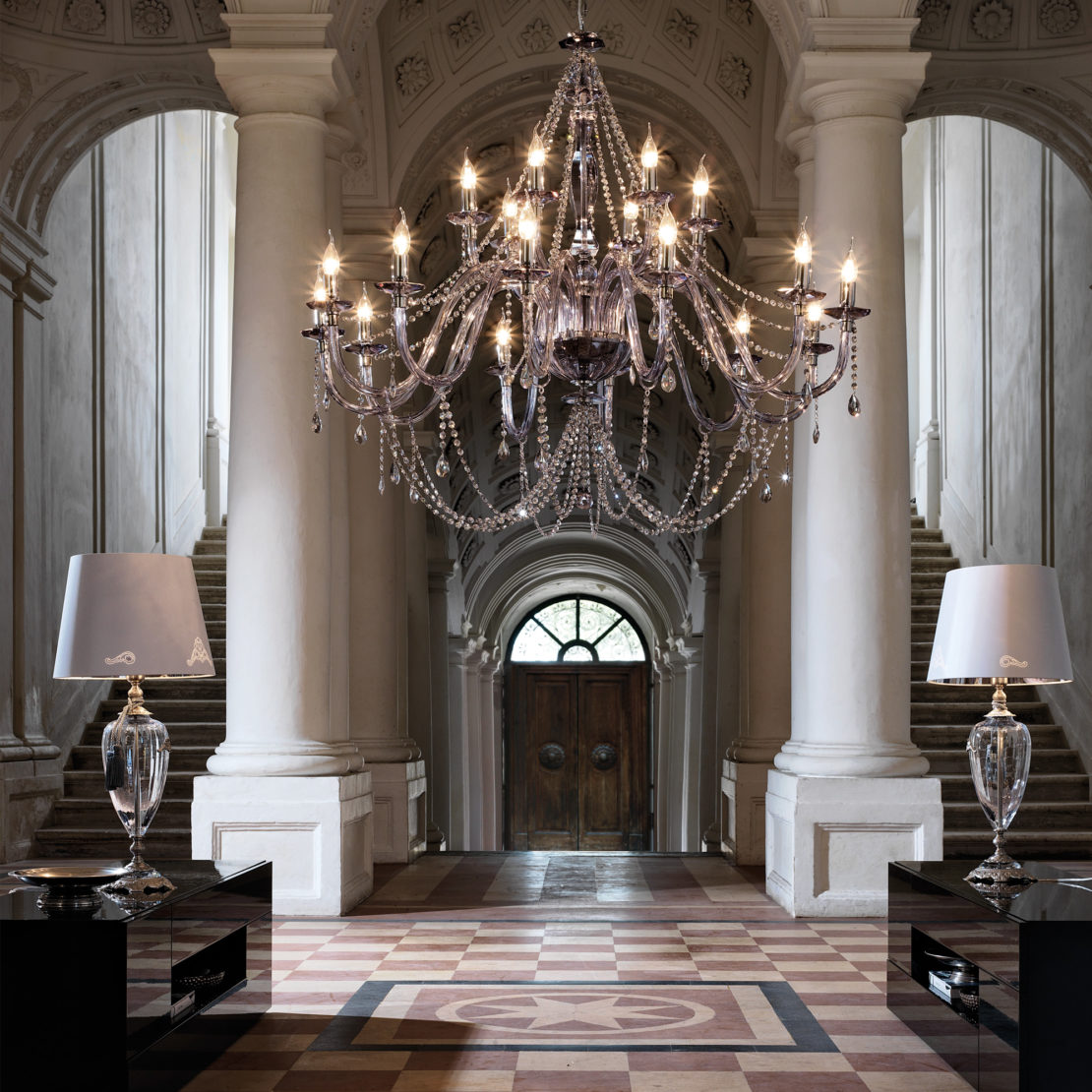 A grand entrance hall with high arched ceilings features the Large Modern Smoked Glass Chandelier, flanked by two staircases. Elegant table lamps sit on black side tables with crystal bases on either side of the checkered floor, leading to a double wooden door at the end.