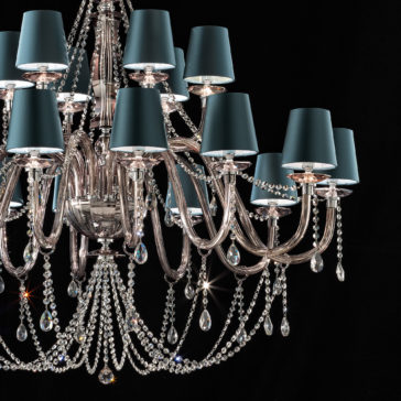 Large Modern Smoked Glass Chandelier With Shades The Large Modern Smoked Glass Chandelier With Shades is an elegant fixture featuring multiple tiers of lights, each enveloped by teal blue lamp shades. This chandelier boasts ornate silver arms and is lavishly adorned with numerous crystal beads and smoked glass pendants, creating a luxurious and sparkling appearance against a dark background.