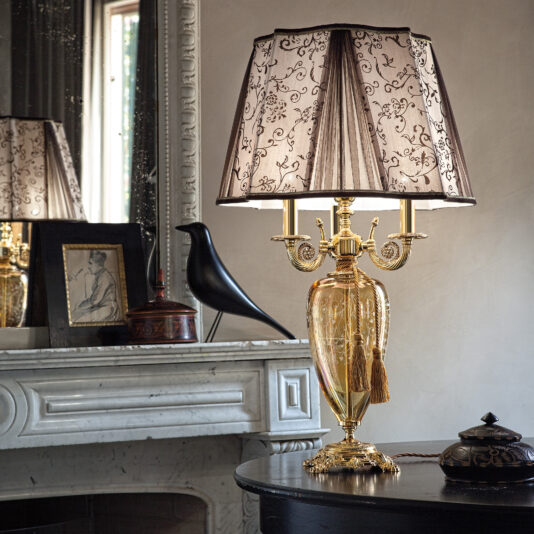 In a chic and sophisticated room, a Large Ornate Amber Glass Table Lamp with a delicately detailed shade graces a dark wooden surface. This elegant lamp stands before an elaborate marble fireplace, complemented by a black bird sculpture and an artful framed picture. The entire scene is reflected in a grand mirror.