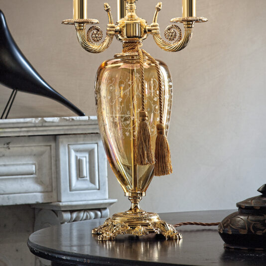 A close-up of the Large Ornate Amber Glass Table Lamp reveals its intricate designs and tasseled details, sitting atop a dark wooden table. In the background, a detailed marble fireplace and a black sculpture enhance the scene, exuding an elegant and vintage aesthetic.