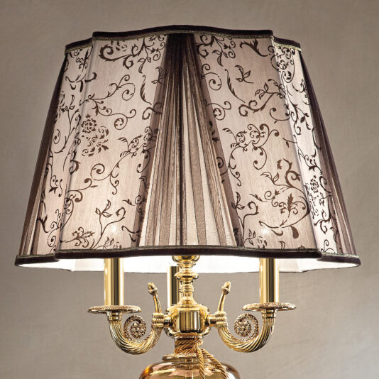 A close-up of the Large Ornate Amber Glass Table Lamp showcases its gold base with intricate scrollwork, complemented by a pleated lampshade adorned with elegant black floral patterns. The lampshade's design adds a touch of classic sophistication to the lamp's overall appearance.