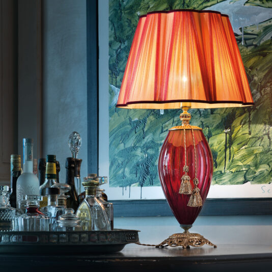 The Large Ruby Blown Glass Table Lamp, featuring an ornate base and a red and gold pleated lampshade, is illuminated and placed on a dark wooden table. Next to it is a silver tray holding an assortment of liquor bottles and a glass decanter, all set against the backdrop of a framed painting.