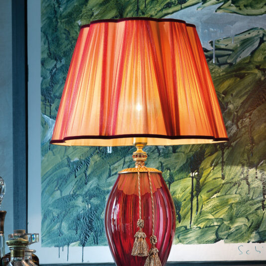 A Large Ruby Blown Glass Table Lamp with a pleated lampshade is showcased against a textured wall adorned with green and beige abstract art. The lamp boasts an intricately designed base and a decorative tassel hanging from the neck, with a small section of a shelf appearing in the background.
