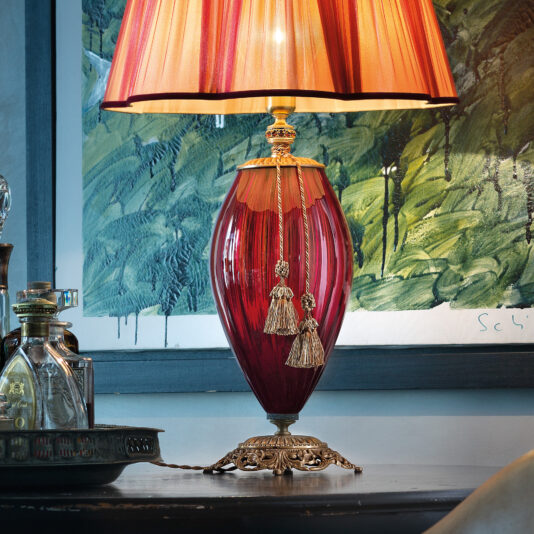 The Large Ruby Blown Glass Table Lamp with gold accents sits on a dark table. The lamp boasts a dual tassel at its base and is topped with a pleated, red and gold lampshade. In the background, a framed green and white abstract painting and glass bottles arranged on a metal tray add to the scene.