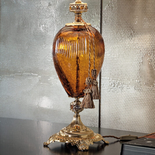 A Large Traditional Amber Glass Table Lamp, adorned with intricate golden metal accents and tassels, is positioned on a dark glossy surface. The lamp is turned off, and the backdrop showcases a textured, metallic-looking wall. An electrical cord leads to a power switch at the base of the lamp.
