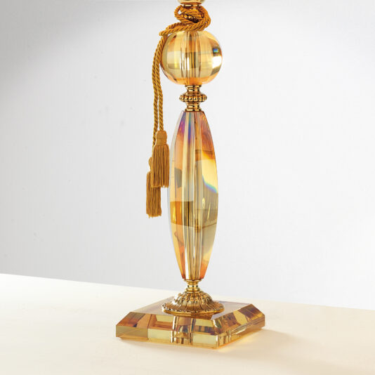 Luxury Amber Glass Table Lamp The Luxury Amber Glass Table Lamp features an ornate cylindrical base with a distinctive sphere near the top and is adorned with decorative tassels. Its reflective, multifaceted design catches light beautifully, creating shimmering effects on its surface. The lamp stands elegantly on a square base.