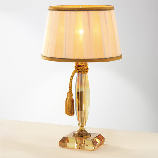 Luxury Amber Glass Table Lamp Introducing the Luxury Amber Glass Table Lamp: A decorative luxury table lamp featuring a golden tassel. This exquisite lamp boasts a translucent, tapered amber glass base and a lampshade adorned with a golden fringe. The shade emits a warm, soft glow, highlighting the sophisticated and elegant design.